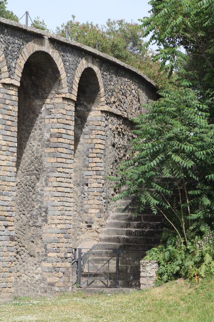 A stone-built viaduct with multiple arches stretches across a garden scene. The arches are constructed from a combination of bricks and flint stones, showing evidence of weathering and age. To the rig