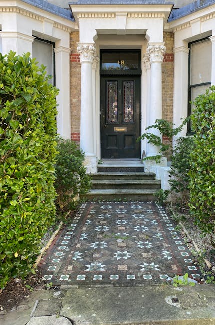A close-up view of the entrance to a period townhouse featuring a black front door with decorative stained glass panels, framed by white classical columns supporting a small overhang. The gray stone s